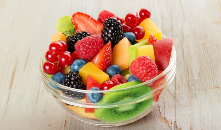 Na foto há um bowl de vidro redondo com diversos tipos de frutas dentro, como kiwi, mirtilo, amora, morango, cerejas e pedaços de manga.