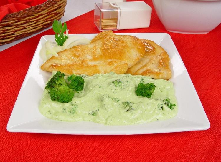 Na foto, a receita está disposta em um prato quadrado de vidro branco, em uma mesa com toalha vermelha. O frango grelhado com creme de brócolis está bem dourado e decorado.