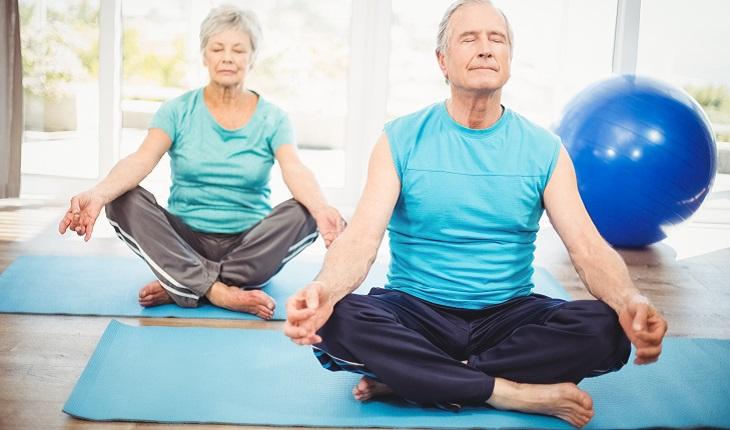 Na imagem, um casal de idosos pratica meditação. Ambos estão com camisetas azuis e sentados. O senhor está na frente e a senhora atrás