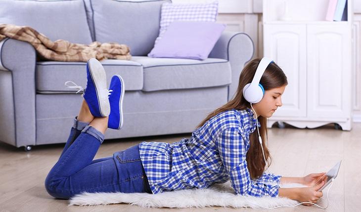 A foto mostra uma menina com cabelo preto e camisa xadrez deitada ouvindo música. Ela está de barriga para baixo no chão e segurando um tablet