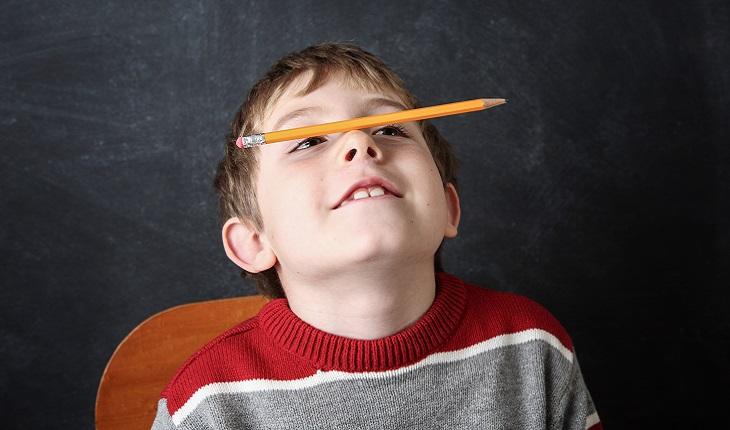 Na imagem há um menino sentado equilibrando um lápis no nariz. Ele está olhando pra cima e sorrindo