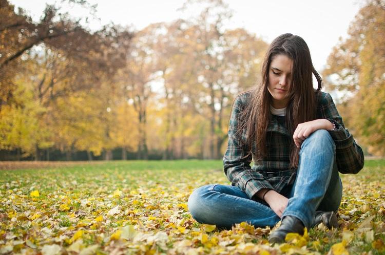 mulher com cara de preocupada sentada na grama de um campo com árvores ao fundo, demonstrando os sinais mais comuns da ansiedade