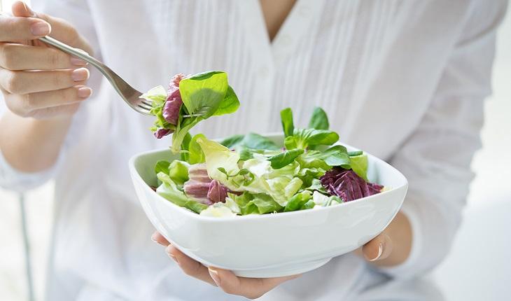 A imagem mostra uma mulher pegando salada de uma tigela com um garfo, visto que a alimentação saudável auxilia na saúde bucal