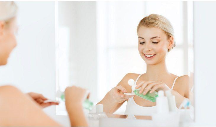 mulher em frente ao espelho passando um produto no rosto com um algodão. Matéria com passo a passo da pele saudável.