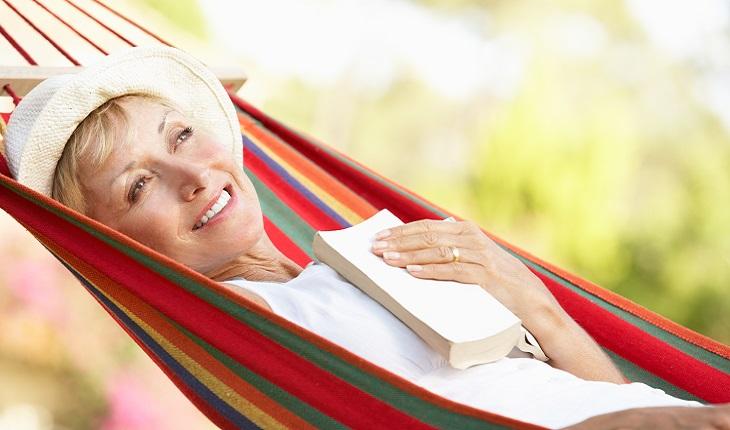 A imagem mostra uma mulher idosa sorrindo com um chapéu deitada em uma rede com um livro encostado no peito.