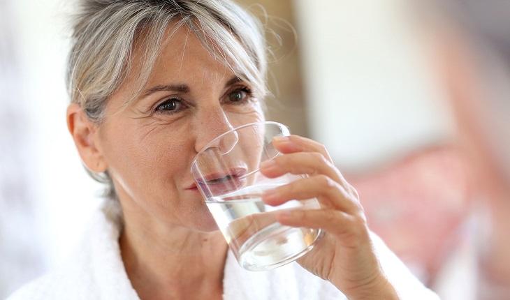 A foto mostra uma mulher idosa com expressão feliz bebendo um copo d'água para melhorar a saúde bucal
