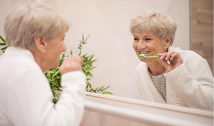 Na foto, é possível observar uma mulher idosa de roupão escovando os dentes em frente a um espelho visando manter a saúde bucal. Ela está com uma expressão contente no rosto