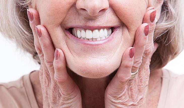 A foto mostra o rosto de uma mulher idosa do nariz para bauxo. Ela está com as mãos nas bochechas e sorrindo, mostrando os dentes muito brancos. A saúde bucal dela está em dia