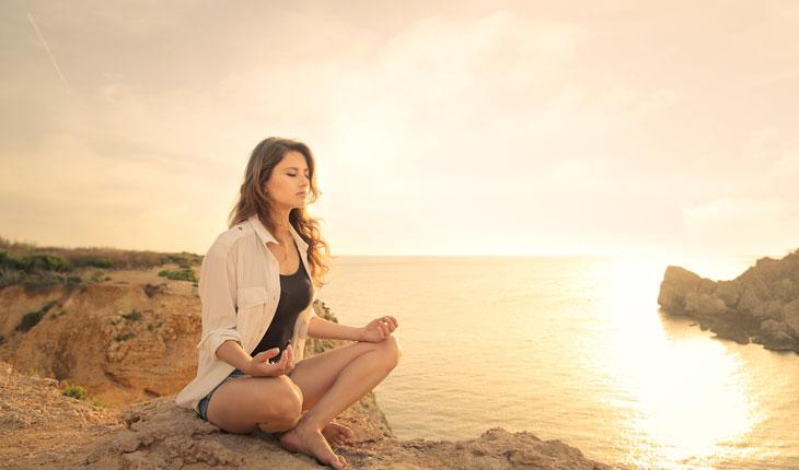 mulher meditando sentada em rochas próximas ao mar ao pôr do sol