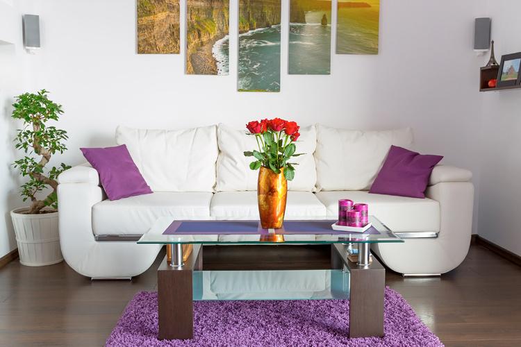 Sala de uma casa decorada com sofá branco e detalhes das almofadas e tapete em roxo. Quadros e plantas para complementar a decoração