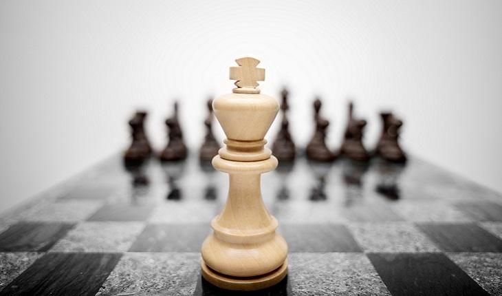 Na imagem, há um tabuleiro de xadrez. A foto foca em um rei bege e ao fundo oito peças pretas desfocadas