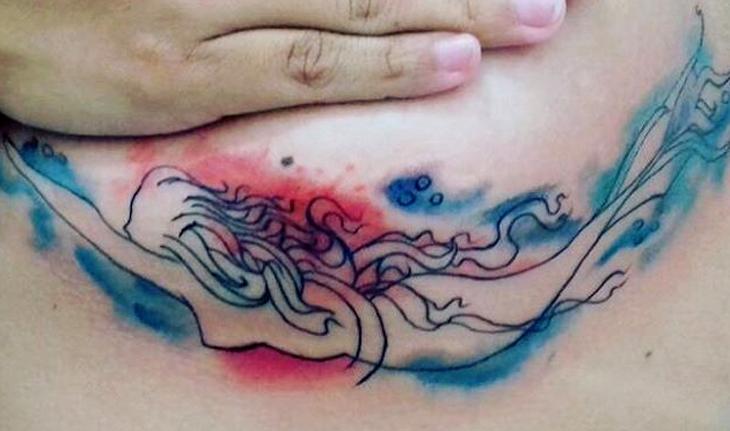 Imagem de tatuagem abaixo do peito de uma sereia nadando com traços simples e toques de aquarela nas cores azul e vermelho ao redor do desenho