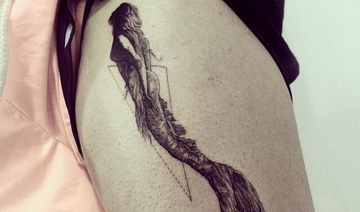 Imagem de tatuagem em preto e branco de uma sereia nadando em direção à superfície com o rosto e busto na sombra e um triângulo com uma das faces tracejada atrás da figura