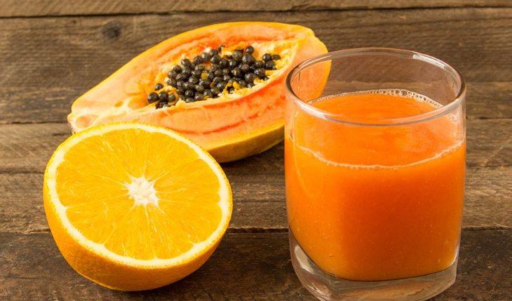 Na foto há um copo com vitamina na cor laranja e um pedaço de laranja e outro de mamão ao lado