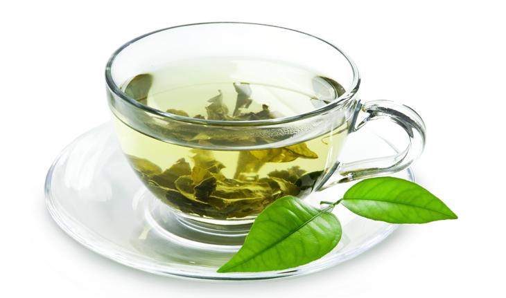xícara transparente com chá verde, ervas no fundo da xícara e folhas verdes decorando