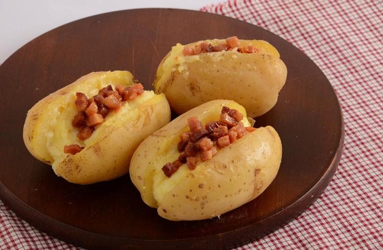 Na foto, a batata recheada com queijo e bacon está em disposta uma tábua marrom. Há três batatas bastante recheadas e polvilhadas com bacon.