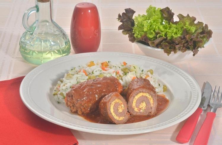 Na foto, o bife rolê a cavalo está em um prato de vidro branco. Nele estão 3 bifes com molho vermelho, acompanhados de arroz com legumes. Na decoração, ao fundo, está uma salada de alface roxa.
