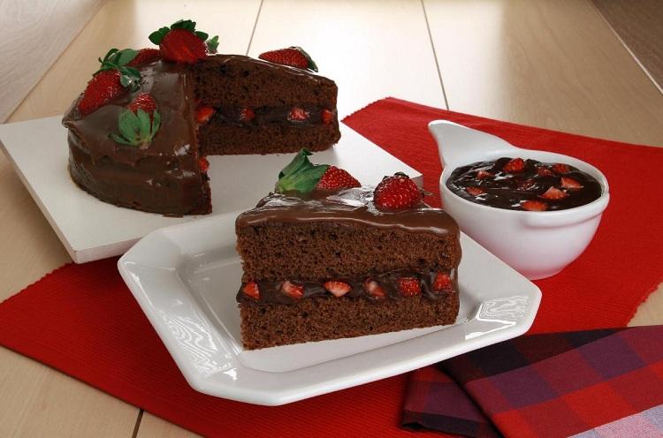 Na foto, o bolo de chocolate com ganache e morango está em um prato de vidro branco. Ao lado, está uma fatia do bolo que mostra uma textura macia com recheio e cobertura cremosos. O bolo está decorado com morangos.