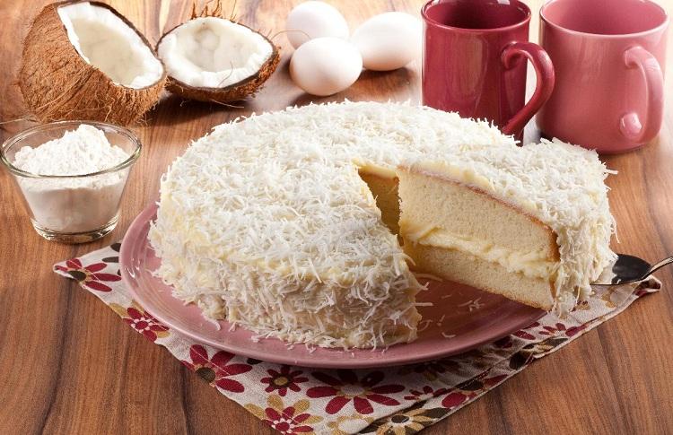 Na foto, o bolo de coco recheado está em uma prato redondo. O bolo tem um pedaço cortado que revela o recheio com creme de coco. Na decoração estão alguns cocos quebrados ao meio e uma tigela com coco ralado.
