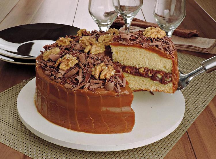 O bolo de nozes recheado tem formato redondo e está sobre um prato branco e um pedaço está sendo cortado, mostrando o recheio de chocolate e nozes