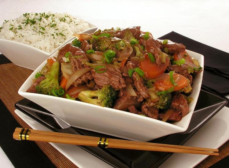 Na foto, a receita de carne com legumes e shoyu está disposta em uma tigela quadrada de vidro branco. Ao fundo, em outra tigela, está arroz branco. Toda a decoração da foto tem inspiração oriental, nas cores branca, preta e marrom.