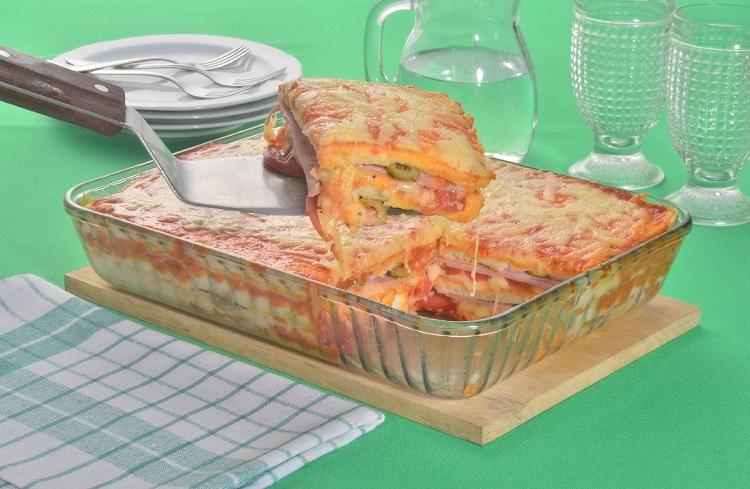 Na foto, a lasanha de pão de forma está em um refratário quadrado de vidro transparente. Uma espátula retira uma fatia da lasanha, mostrando as camadas de queijo, presunto e molho vermelho. Na decoração ao fundo, estão pratos, copos e uma jarra de vidro.