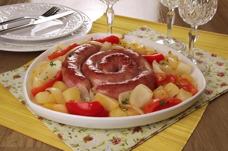 Na foto, a linguiça assada com batatas está uma fôrma redonda, temperada com cebola e tomates fatiados. A receita está bem dourada, e na decoração estão talheres, pratos e taças de vidro.