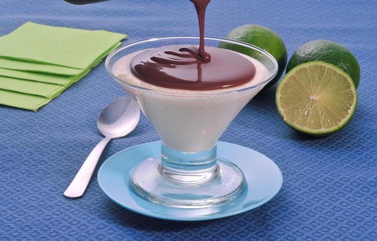 Na foto, o mousse de limão com calda de chocolate está disposto em uma taça pequena de vidro. A calda de chocolate está sendo despejada sobre o mousse. A mesa está coberta com uma toalha azul e na decoração está uma colher e limões fatiados ao lado.