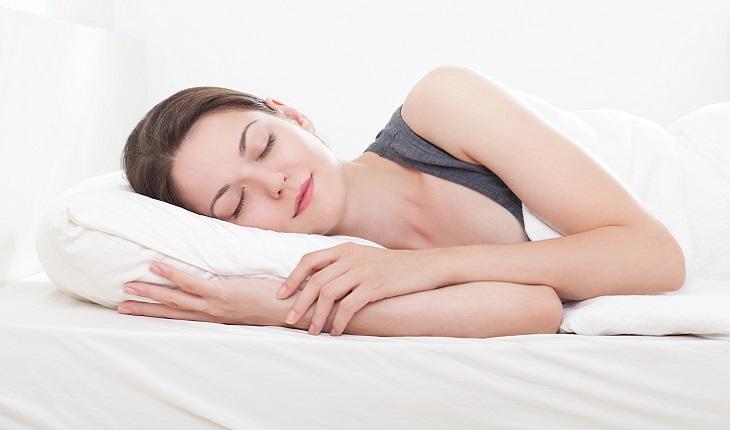 A foto mostra uma mulher com cabelo e camiseta pretos dormindo. Ela está deitada em uma cama branca