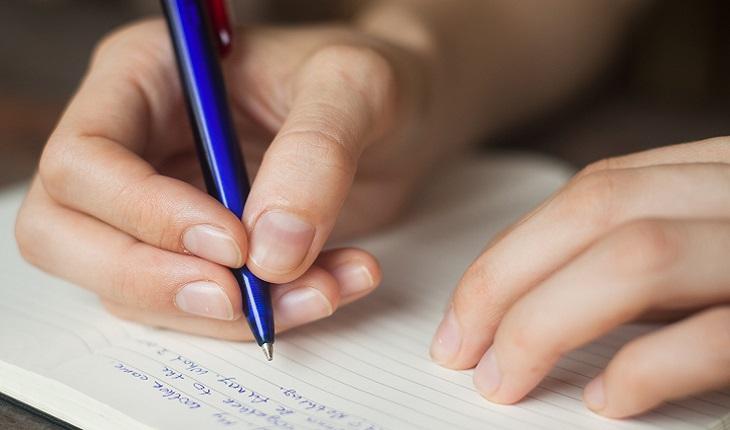 A foto mostra as mãos de uma mulher. Ela está escrevendo com uma caneta azul em um caderno
