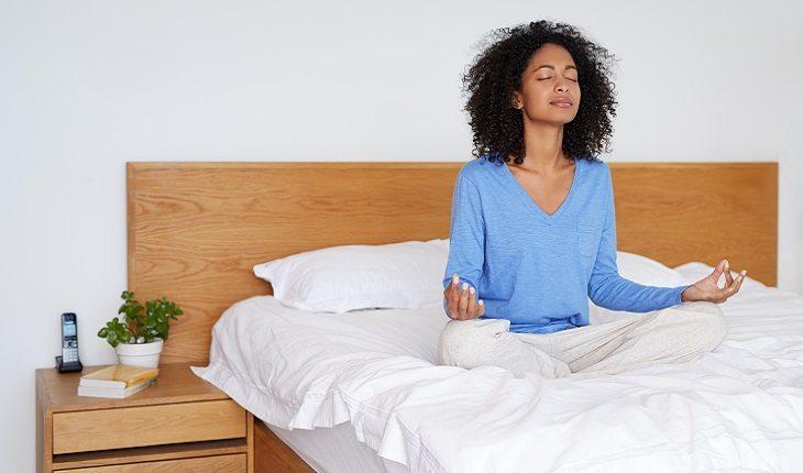 A foto mostra uma mulher sentada na cama meditando