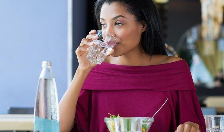A imagem mostra uma mulher negra com uma camiseta vermelha bebendo um copo de água. Ela está com uma expressão tranquila. A sua frente ainda há uma tigela com salada e uma garrafa com água