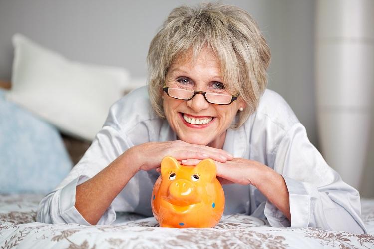 mulher grisalha sorrindo com o rosto e as mãos apoiados em um cofre-porquinho laranjado, organizar os gastos