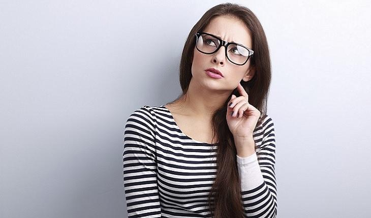 A foto mostra uma mulher com cabelo preto aparentando uma expressão de curiosidade. Ela está usando uma camiseta listrada em preto e branco e óculos.