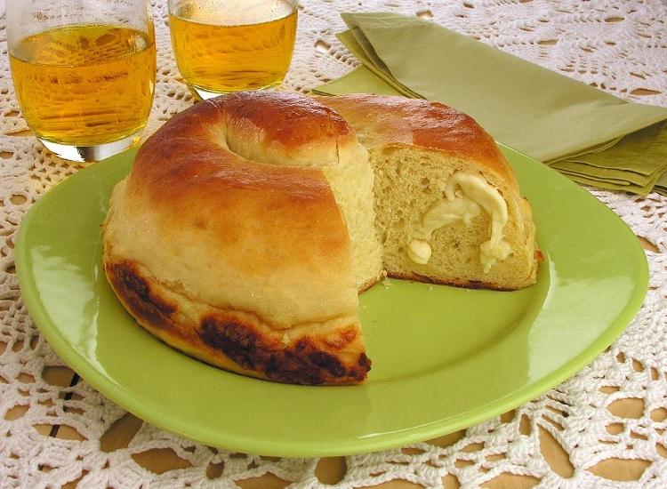 Na foto, o pão de mandioca com queijo está em um prato de vidro verde. O pão tem uma fatia cortada, revelando uma massa macia com pedaços de queijo. Ao fundo estão dois copos com refrigerante.