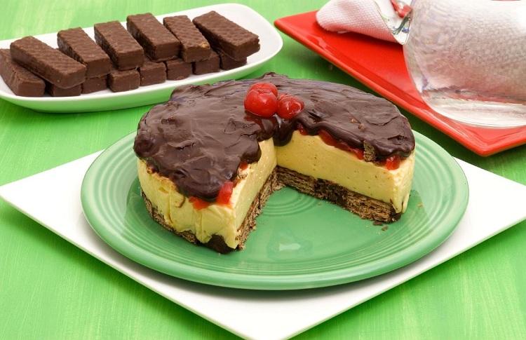 Na foto, a torta Bis® com cereja está disposta em um prato verde de vidro. A torta é redonda e está cortada, revelando a textura das camadas. Na decoração, ao fundo, está uma travessa com chocolates Bis® e um prato vermelho.
