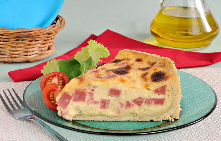Na foto, um pedaço triangular da torta de queijo com mortadela sobre um prato de vidro e acompanhado de alface e tomate-cereja 