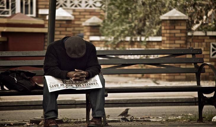 Homem negro sentado em ponto de onibus com um jornal na mão e cabeça baixa rezando. Provérbios