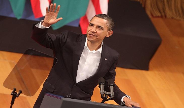 Barack Obama fazendo um discurso e acenando enquanto se posiciona em frente a um microfone