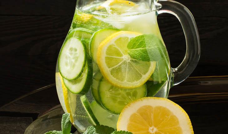 foto de uma jarra de vidro transparente com água saborizada de limão, pepino e hortelã, com pedaços dos ingredientes na jarra e decorando ao redor dela