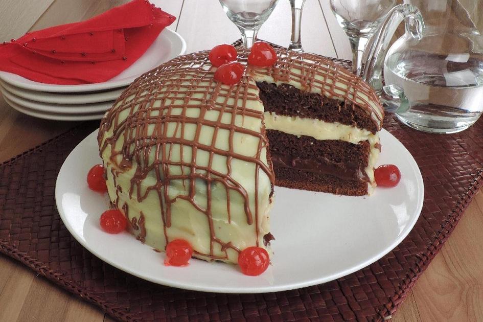 Na foto, o bolo de chocolate branco e preto está disposto em um prato de servir. O bolo está decorado com fios de chocolate preto e cerejas. Um pedaço do bolo está cortado e foto revela o recheio de duas camadas.