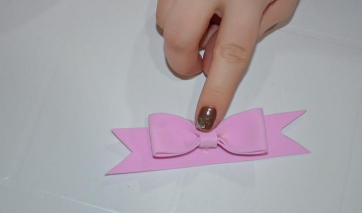 Na foto é possível ver as mãos de uma pessoa colando o laço feito de E.V.A. rosa em uma tira de E.V.A. rosa