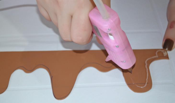 Na foto é possível ver as mão de uma pessoa passando cola quente utilizando uma pistola de cola na cor rosa. A pessoa está passando a cola por um E.V.A. marrom cortado em formato de calda de bolo, com curvas.