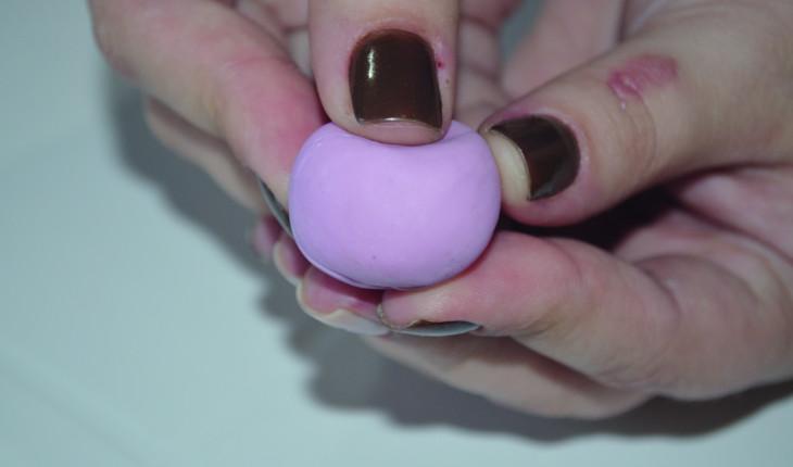 Na foto é possível ver a mão de uma pessoa modelando a massa de biscuit rosa, fazendo um bolinha e depois achatando a parte de cima.