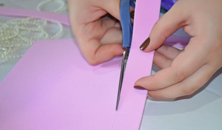 Na foto é possível ver as mãos de uma pessoa cortando uma tira de E.V.A. rosa.