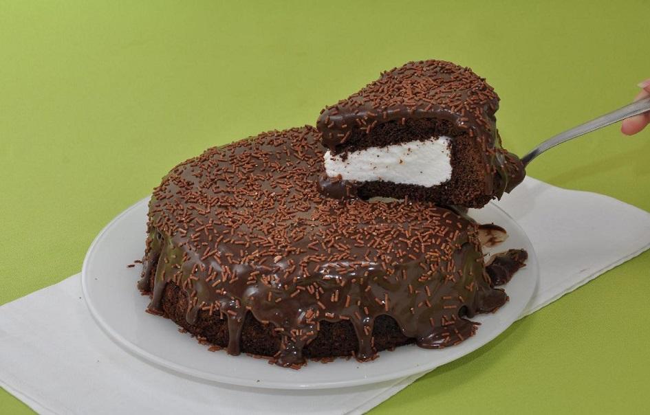 O bolo de marshmallow e brigadeiro é redondo e está sobre um prato branco também redondo. Um pedaço está sendo cortado, sendo possível ver o recheio de marshmallow branco. O bolo também tem cobertura de chocolate e granulado por cima.