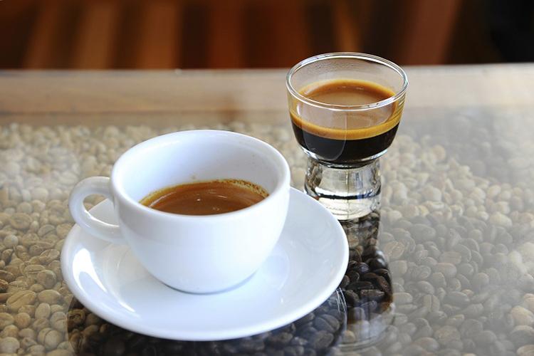 Matéria sobre estudo que aponta que cafeína ajuda na memória. Na foto, Uma xícara branca e copo com café sobre uma mesa com grãos do produto na parte inferior.