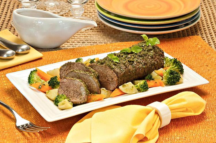 A carne assada com manjericão e legumes está sobre uma travessa branca e os legumes estão ao redor. A travessa está sobre uma mesa com itens de cozinha em tons laranja.