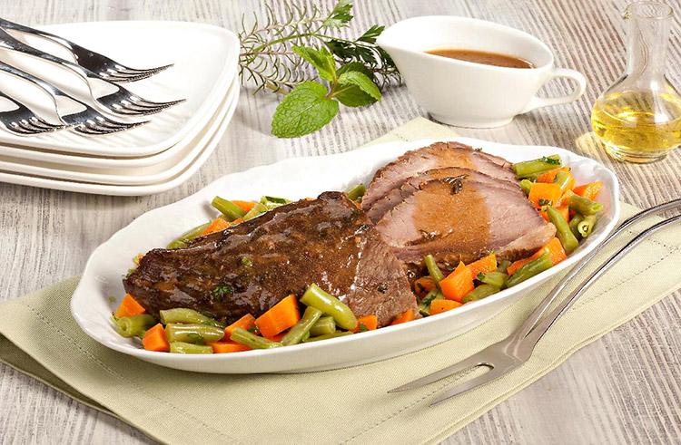 A carne ao forno com ervas está em um prato branco oval e está fatiada em uma parte da peça e há legumes ao redor