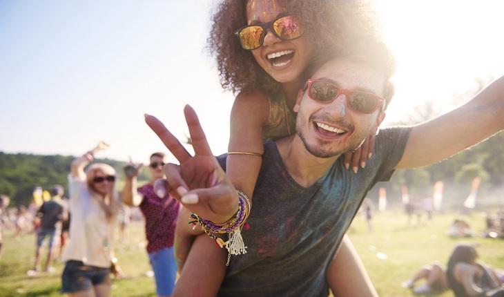 Homem carregando mulher nas costas, sorrindo e fazendo sinal de paz e amor num festival durante o dia
